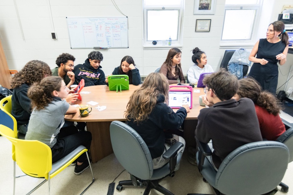Youth sitting around a table with iPads listening to woman teacher speak